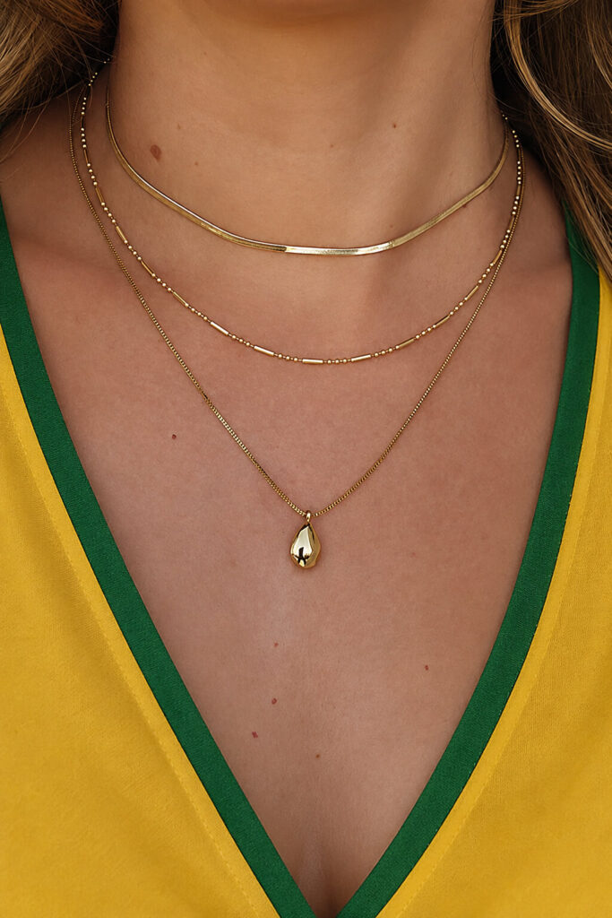 Colo de uma mulher com mix de colares dourados com pingente gota, combinando com blusa amarela e verde em look inspirado na Copa.